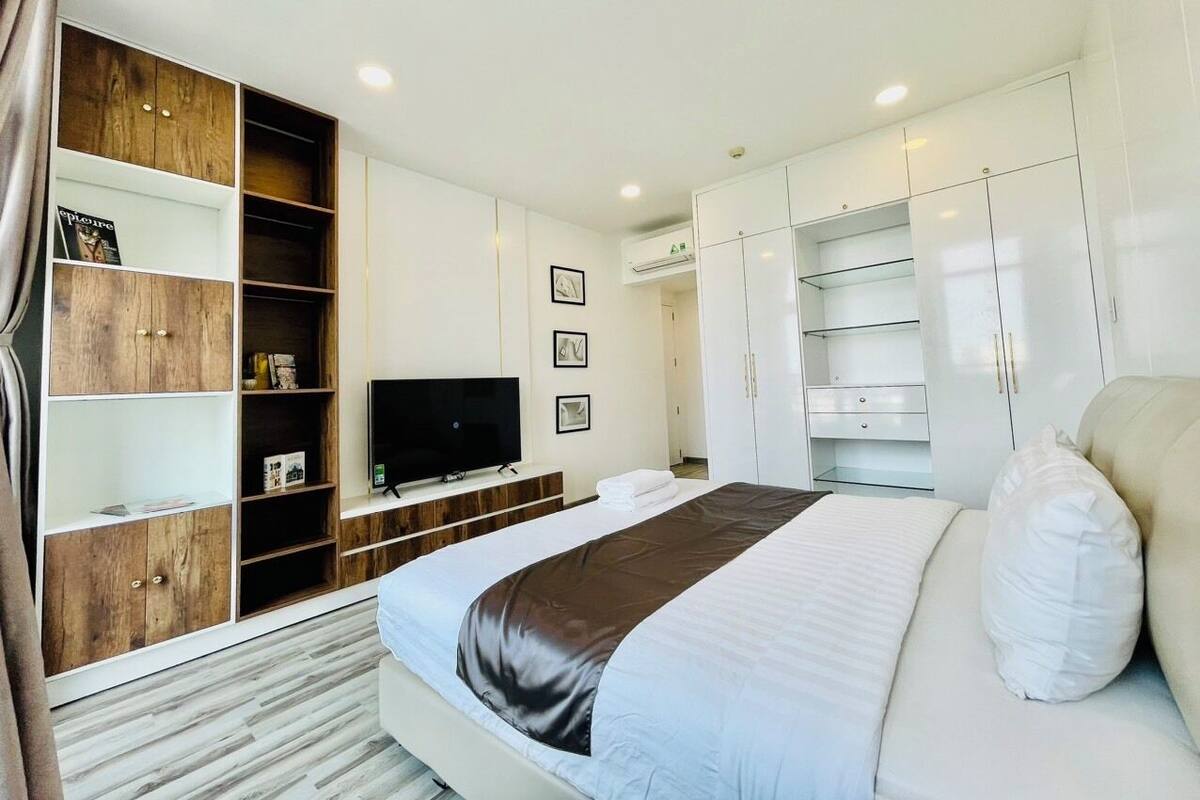 A modern hotel room with a large bed, built-in wardrobe, a flat-screen TV, and a seating area with a sofa and pillows.