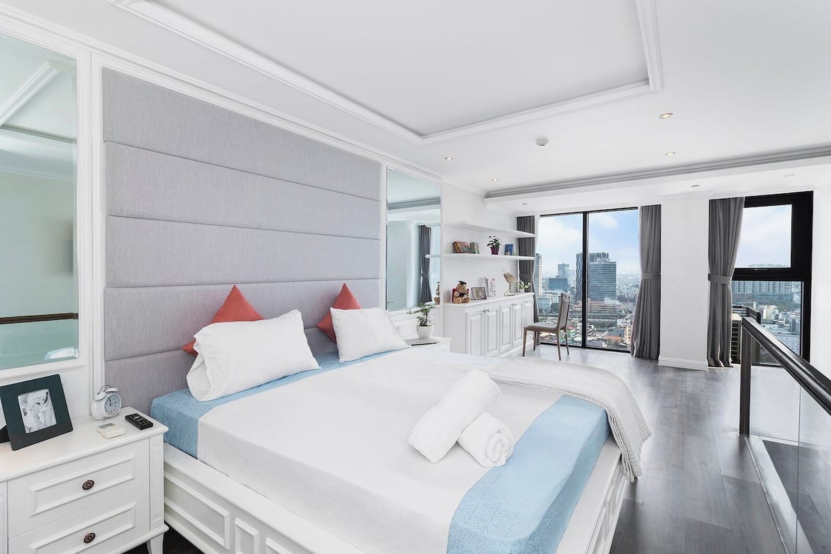 A modern hotel room with a large bed, a nightstand, a dresser, and a view of the city through large windows.