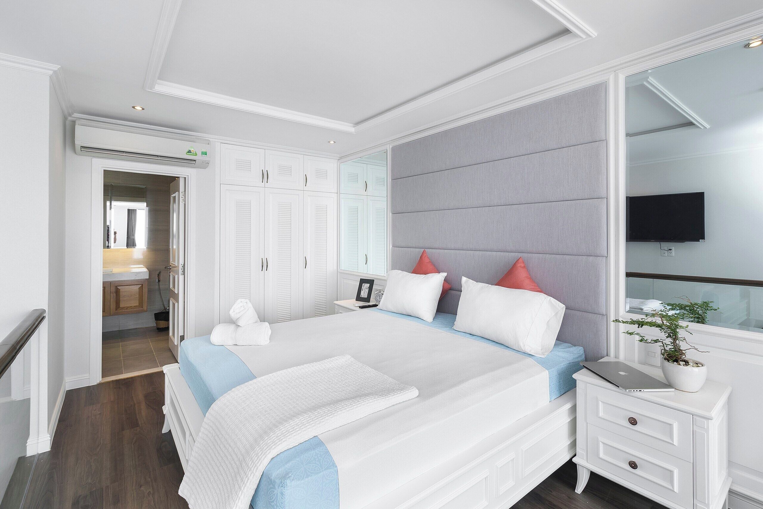 A hotel room with a large bed, a bedside table with a laptop, a potted plant, and a TV mounted on the wall.