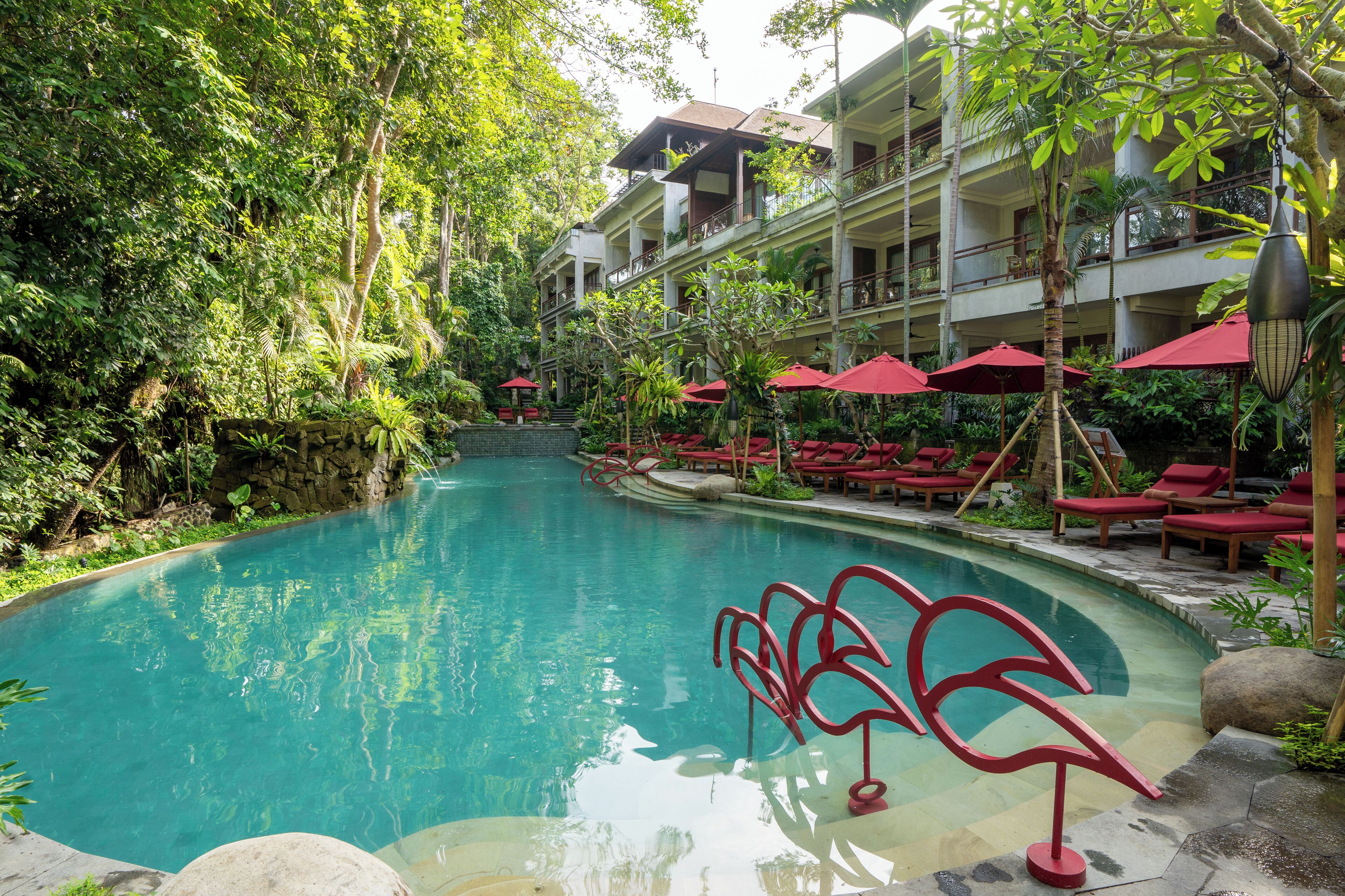 A pool surrounded by lush greenery, with red umbrellas and lounge chairs.