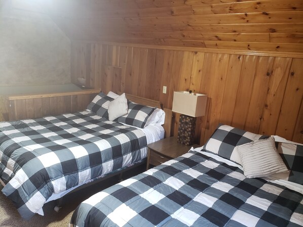 A room with two beds, each with a checkered bedspread, wooden walls, and a small bedside table with a lamp.