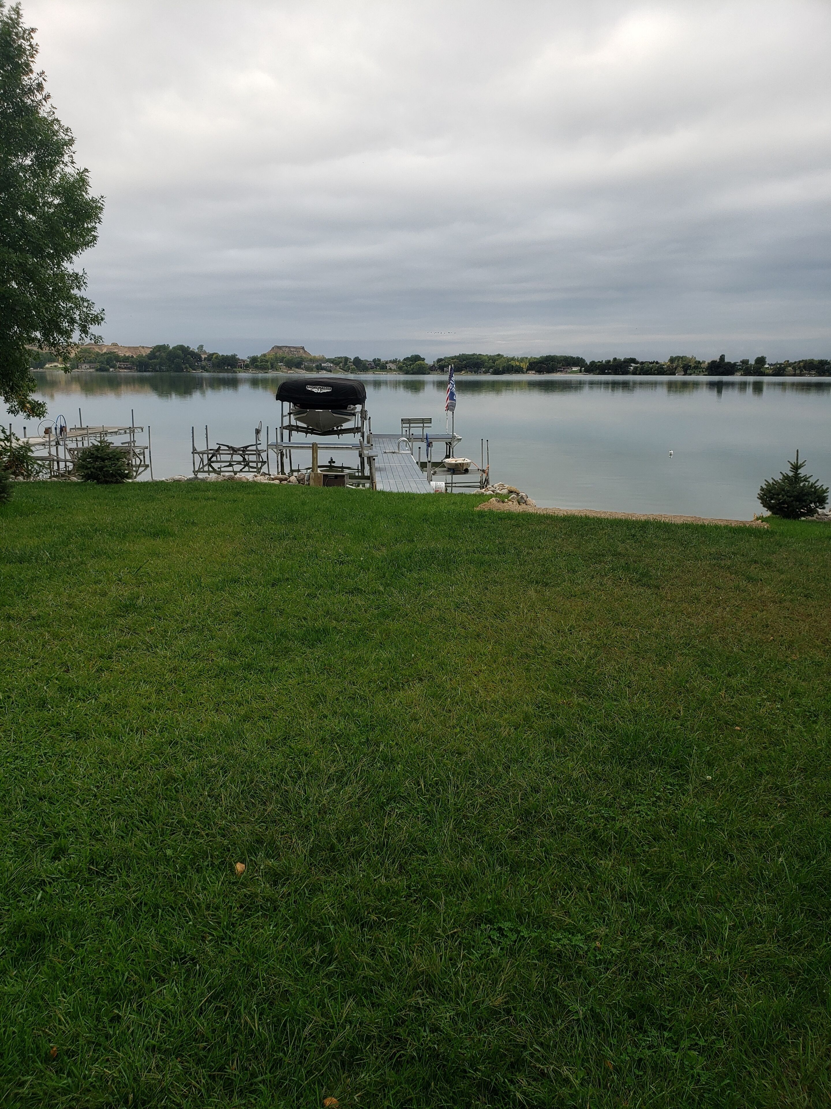 A grassy area with a dock extending into a calm lake, surrounded by trees and hills in the distance.