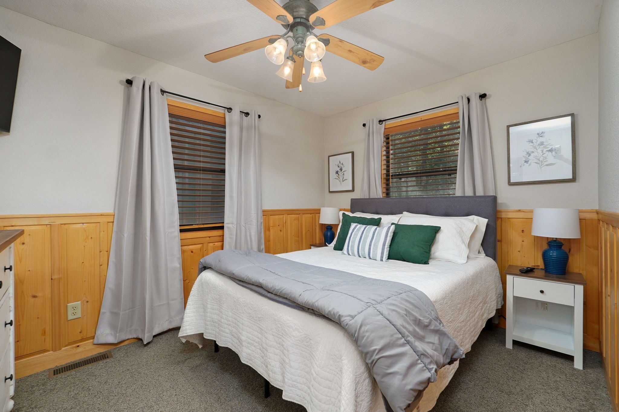 A bedroom with a bed, wooden headboard, two bedside tables, a ceiling fan, and a window with blinds.
