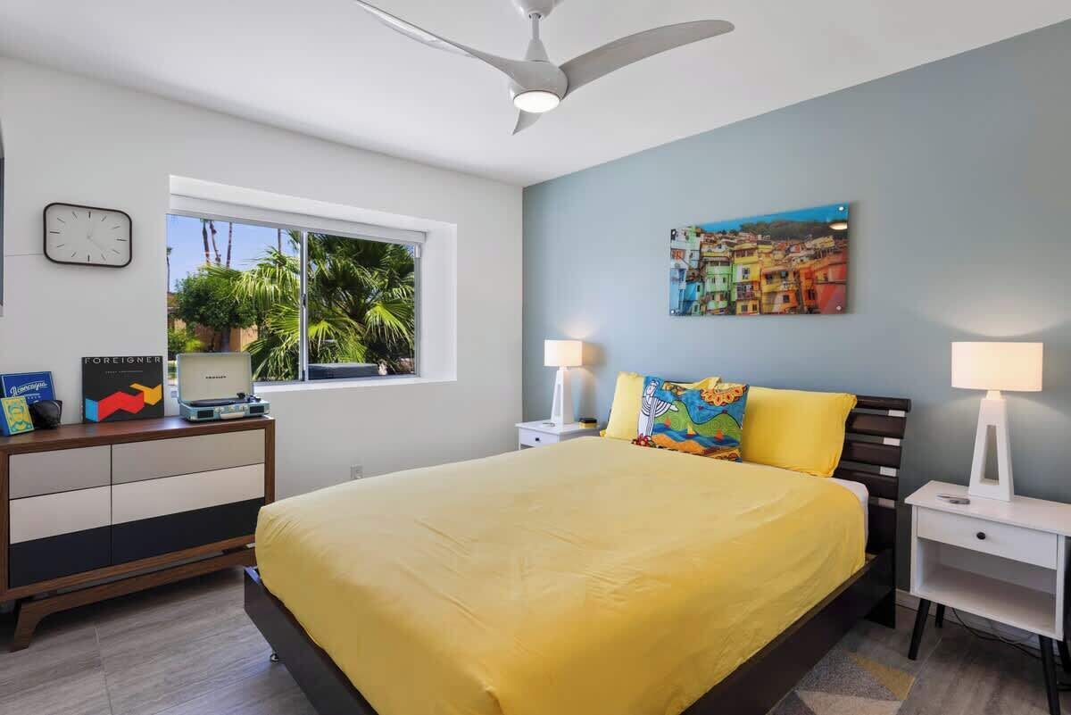 A bedroom with a bed, a desk, a chair, a lamp, a clock, and a window with a view of palm trees.