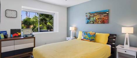 A bedroom with a bed, a desk, a chair, a lamp, a clock, and a window with a view of palm trees.