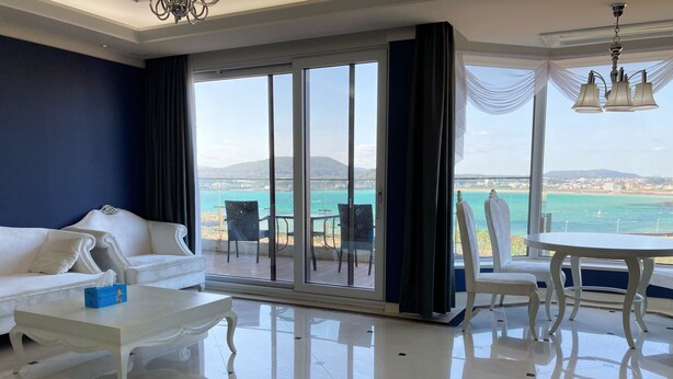 Una sala de estar moderna con un sofá blanco, una mesita de centro y un comedor con mesa y sillas. Amplias ventanas ofrecen vistas al mar y un horizonte urbano distante.