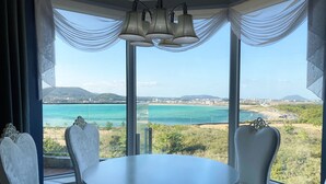 Un comedor con una mesa redonda y sillas blancas, ventanas amplias con cortinas translúcidas y vistas a un paisaje costero.