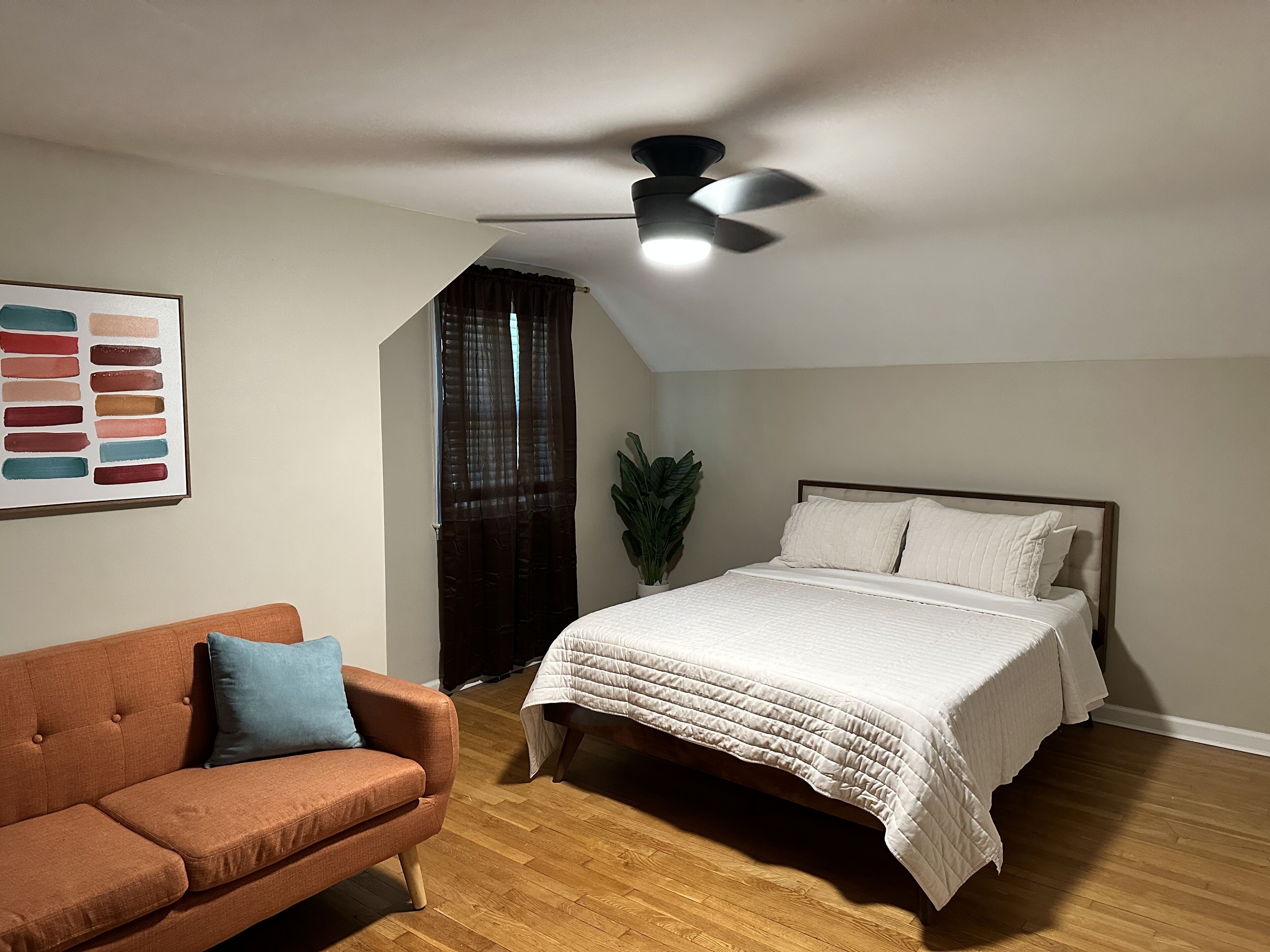 A bedroom with a bed, a ceiling fan, a brown sofa, a potted plant, and a wall with a color palette.