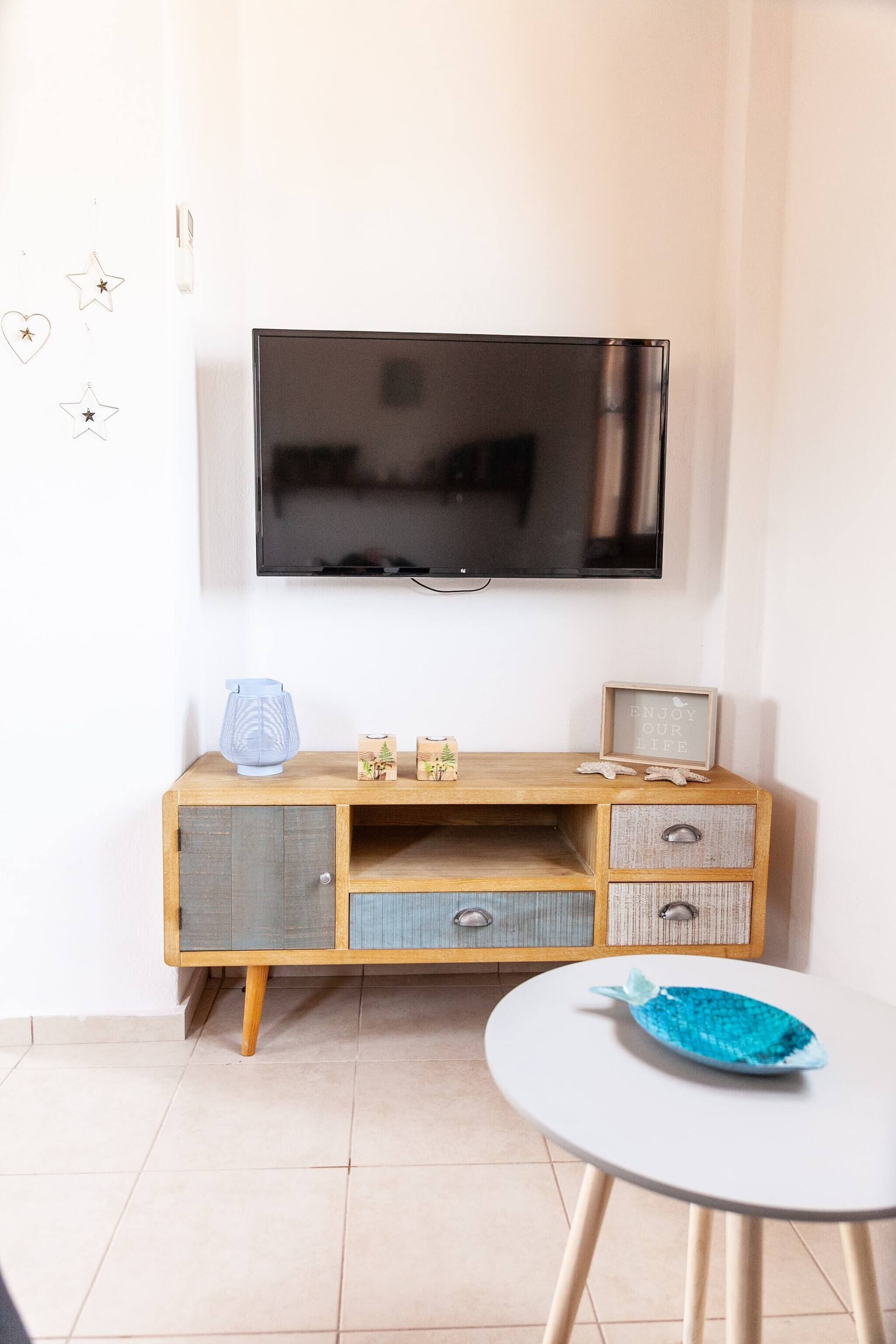 A room with a TV mounted on the wall, a wooden cabinet with drawers, a small table, and a decorative fish on the table.