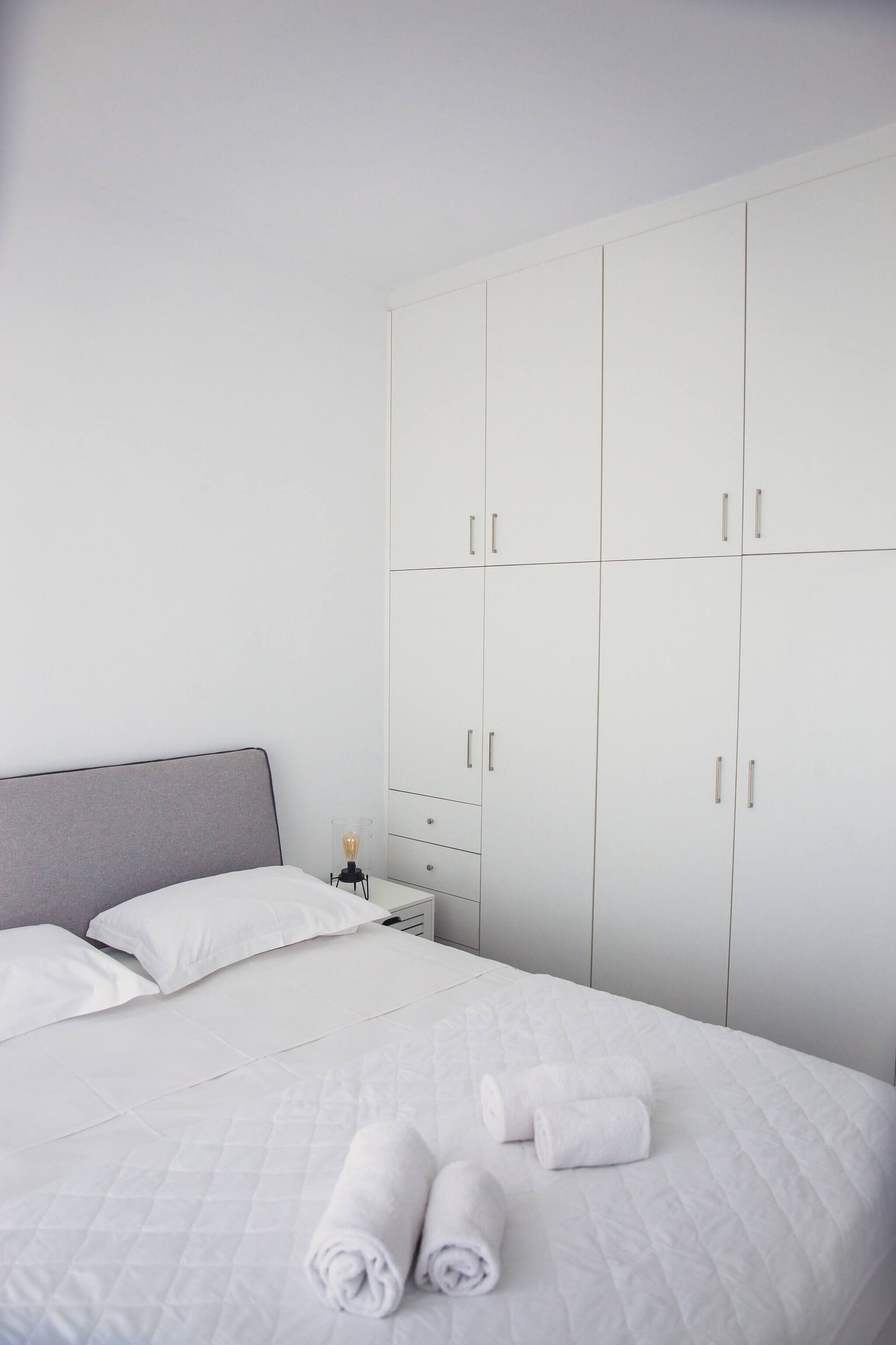 Een keurig opgemaakt bed met witte lakens en opgerolde handdoeken, een bedlamp en een grote witte garderobe met meerdere deuren.