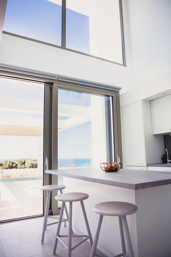 A modern kitchen with a large window, two stools, and a fruit bowl on the counter.