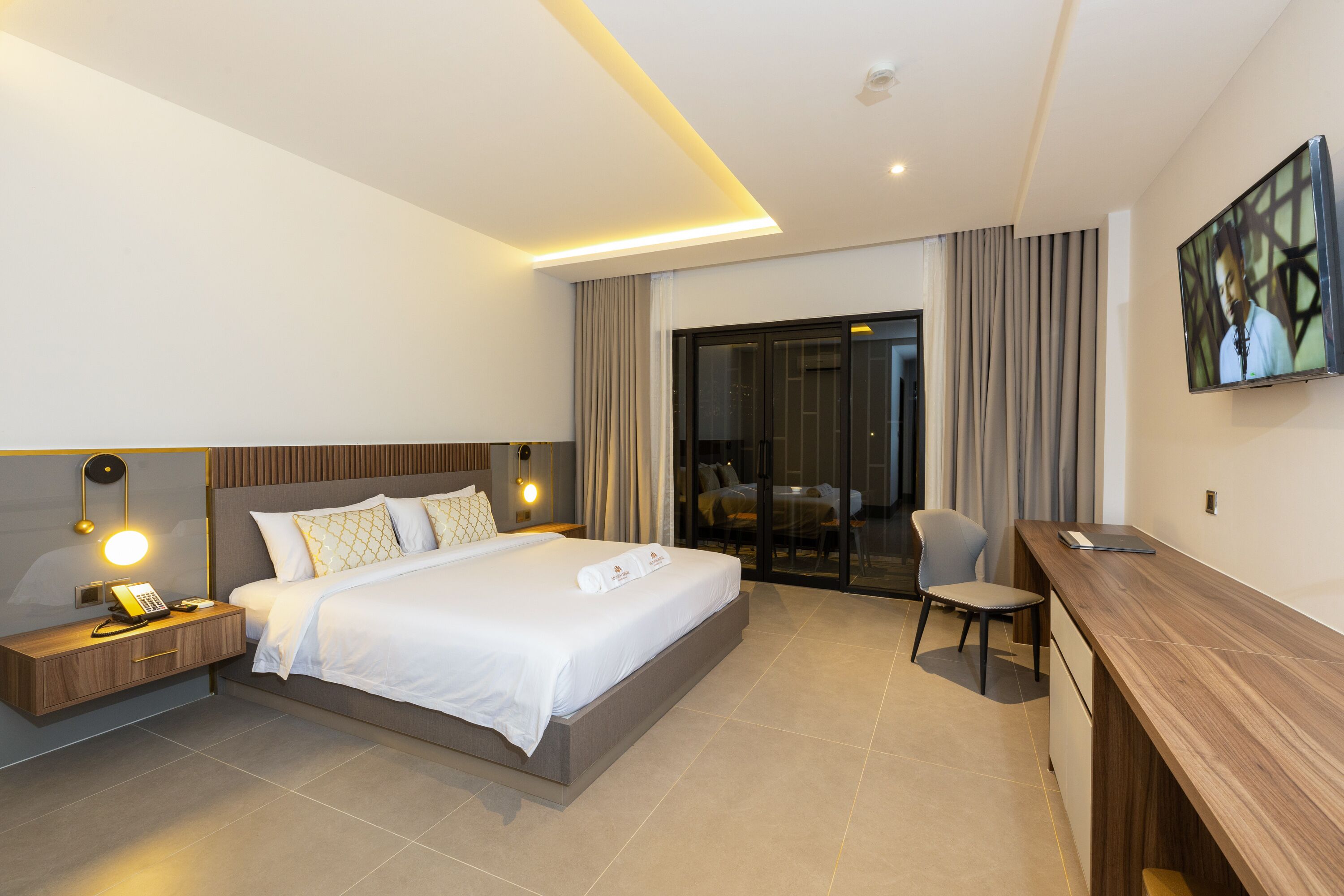 A modern hotel room with a large bed, bedside table with a lamp, a desk, and a TV.