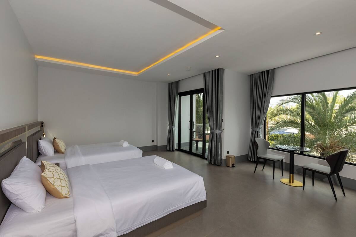 A hotel room with two beds, a dining table with chairs, and a large window with a view of palm trees.