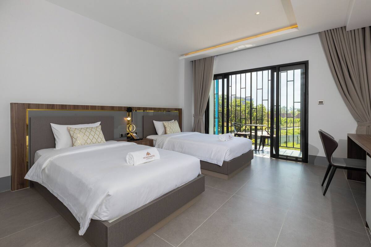 A hotel room with two beds, a desk, and a balcony with a view.