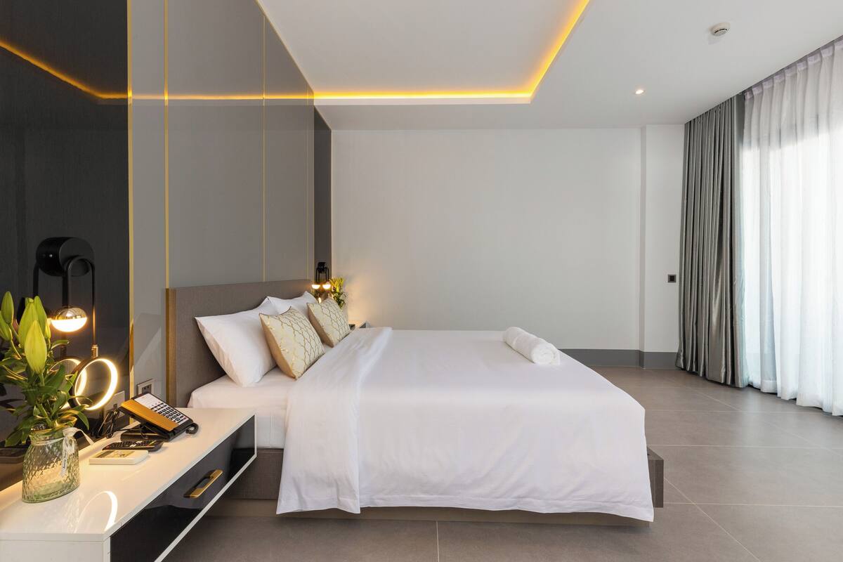 A modern bedroom with a large bed, bedside tables, a desk with a lamp, and a large window with curtains.