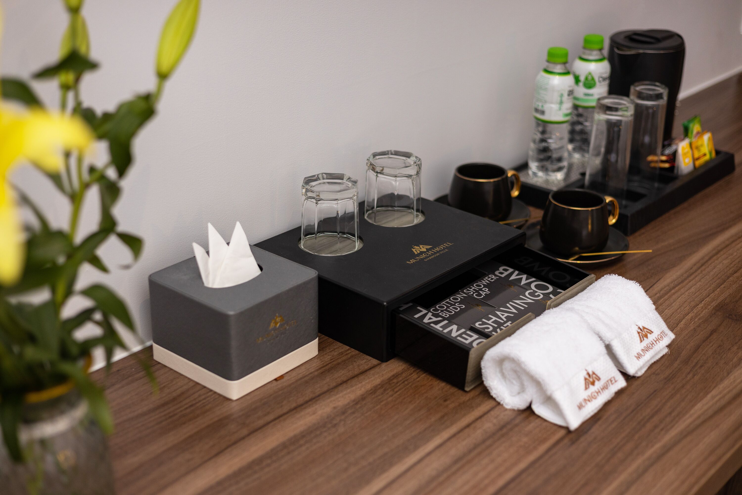 A neatly arranged hotel room with a tissue box, two glasses, a black tray with bottles and a coffee maker, and a rolled towel.