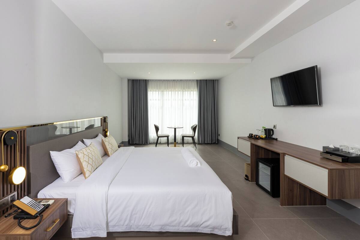 A modern hotel room with a large bed, bedside table with a lamp, a desk with a flat-screen TV, and a small seating area with a table and chairs.