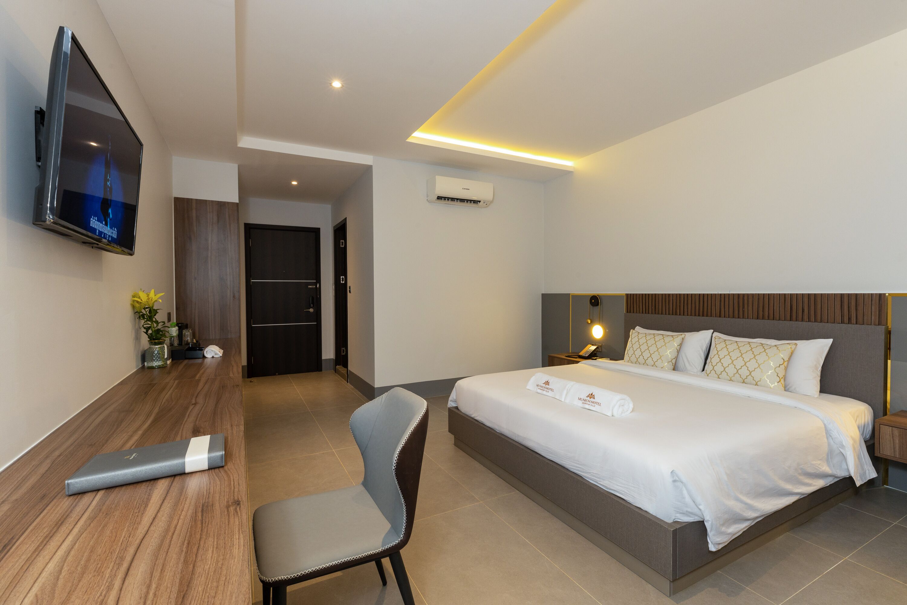 A modern hotel room with a large bed, a desk with a laptop, a TV mounted on the wall, and a small plant on the desk.