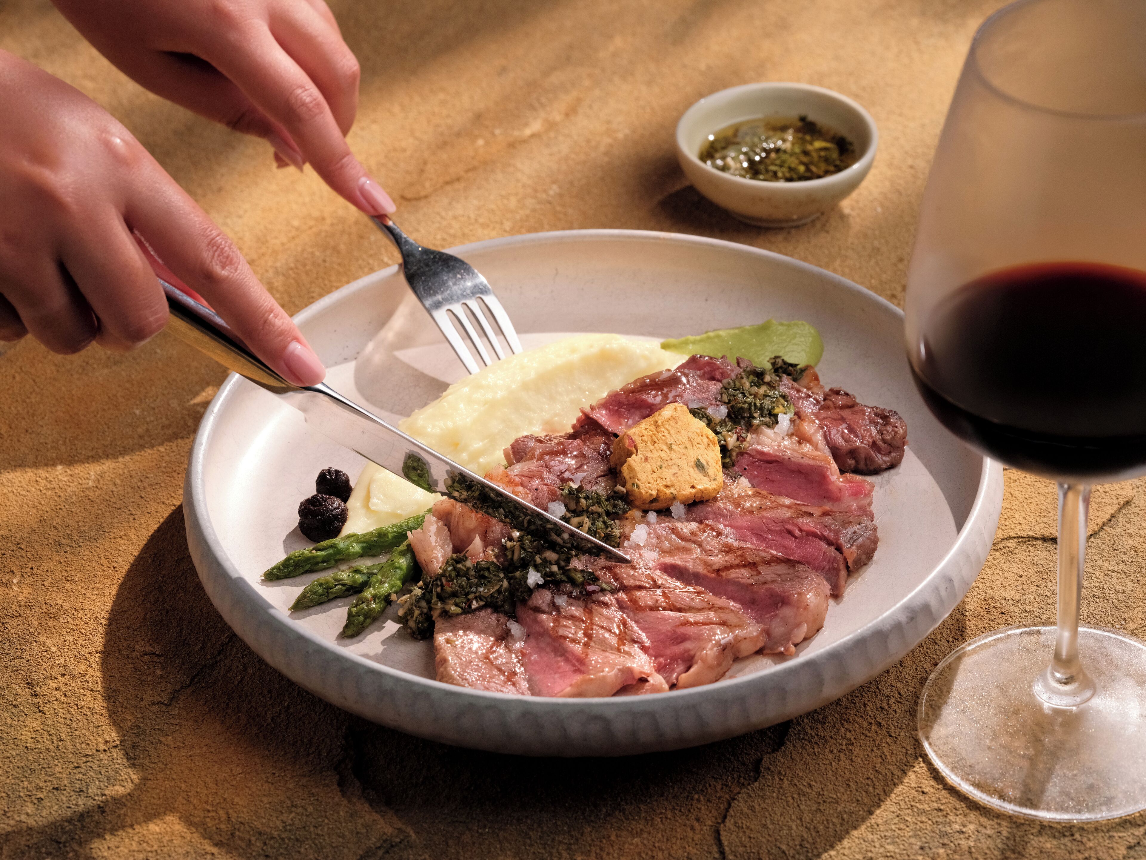 Un plato de comida con un tenedor y cuchillo, una copa de vino tinto y un recipiente pequeño con salsa sobre una mesa.