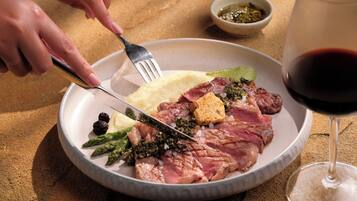 Un plato de comida con un tenedor y cuchillo, una copa de vino tinto y un recipiente pequeño con salsa sobre una mesa.