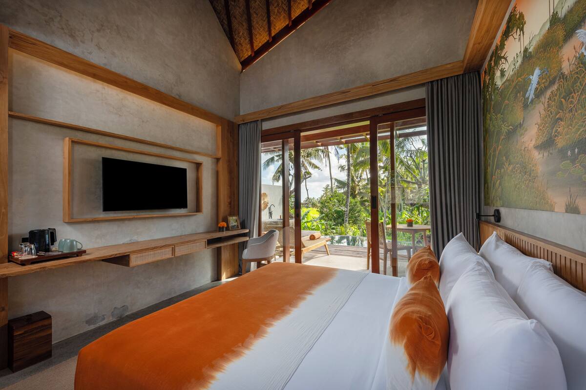 A hotel room with a large bed, a flat-screen TV mounted on the wall, a wooden desk with a chair, and a view of palm trees through the glass door.
