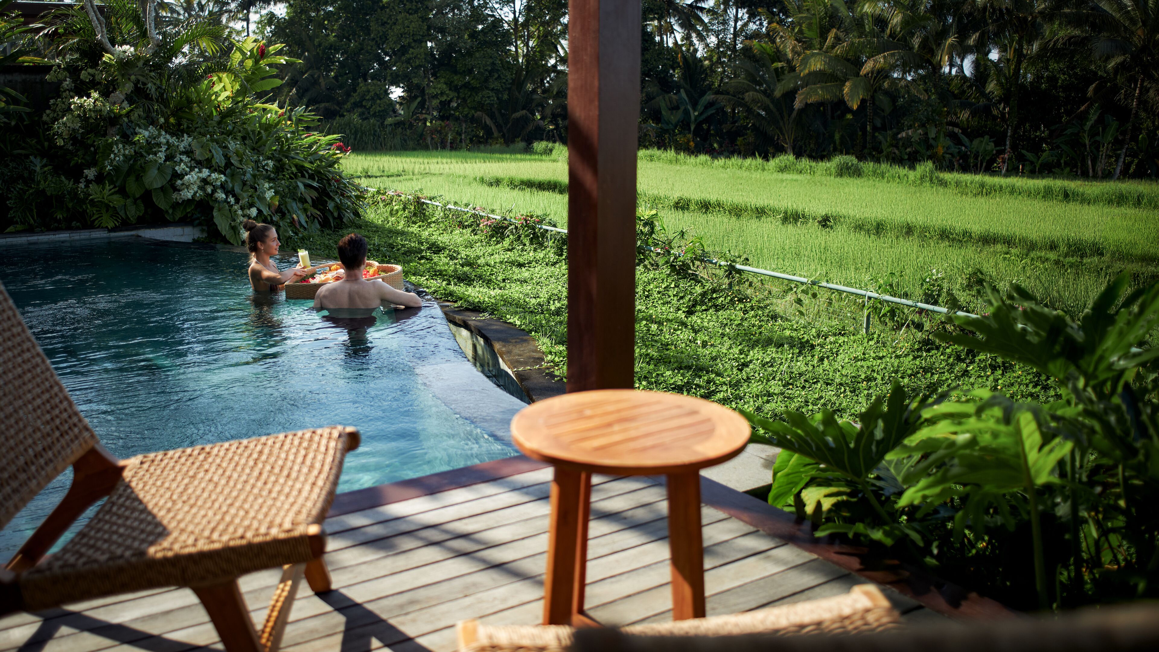 A pool area with two wicker chairs and a small wooden table, surrounded by lush greenery and a rice field in the background.