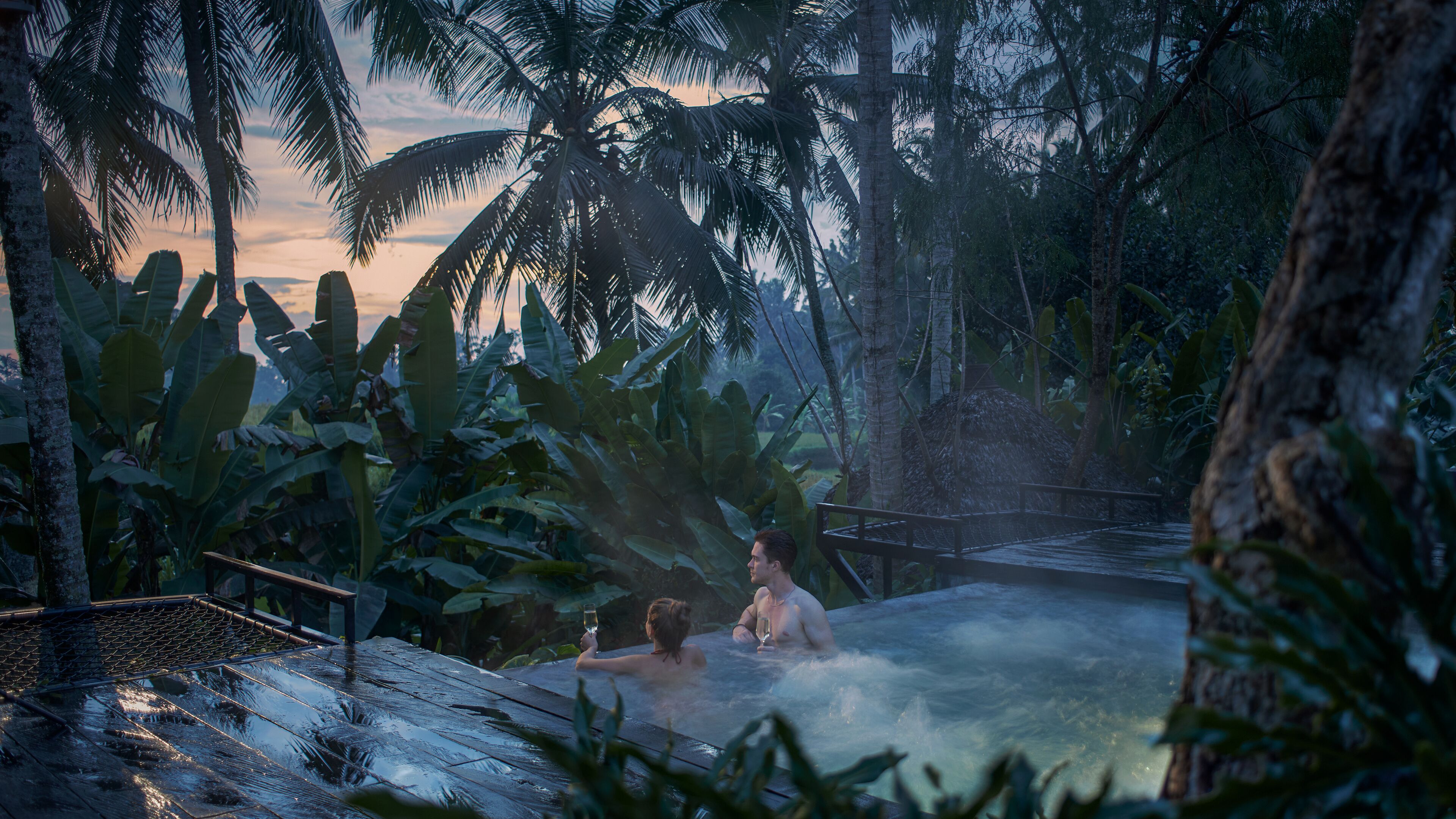 A hot spring area with two people relaxing in the water, surrounded by lush tropical vegetation and a wooden bridge.
