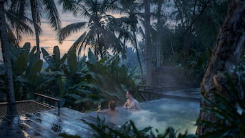A hot spring area with two people relaxing in the water, surrounded by lush tropical vegetation and a wooden bridge.