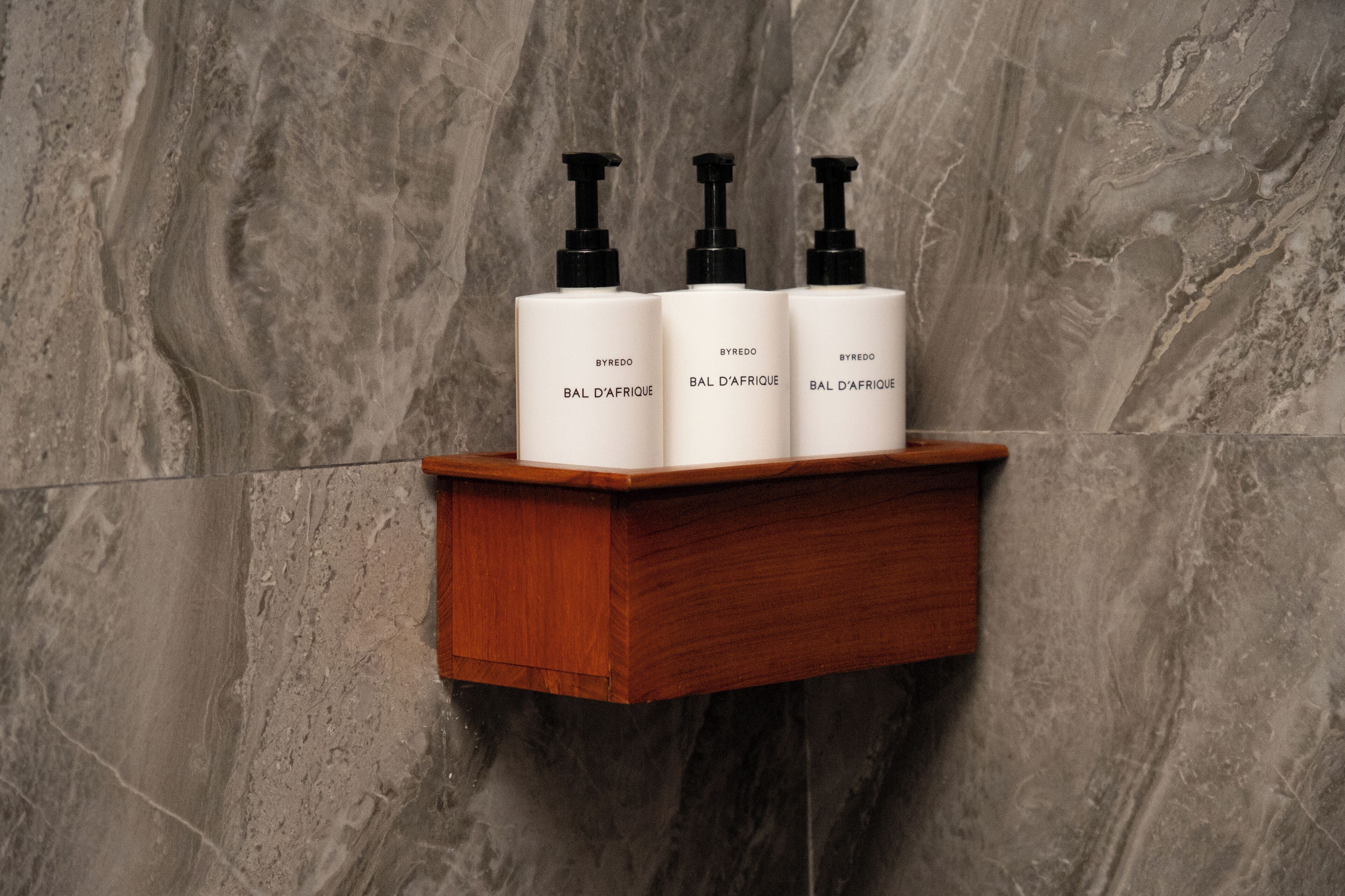 A wooden shelf with three white bottles labeled "Byredo BAL D'AFRIQUE" against a marble wall.
