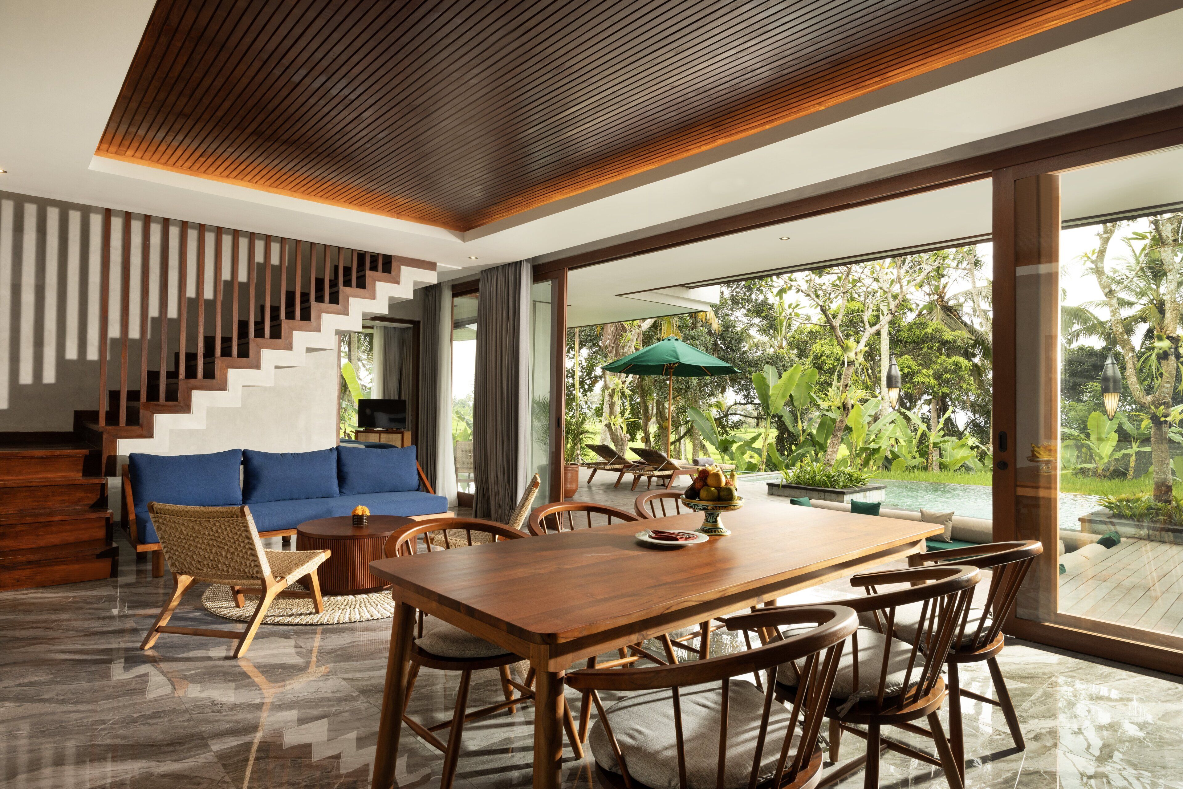 A modern living room with a wooden dining table, blue sofa, and a staircase with wooden railings.