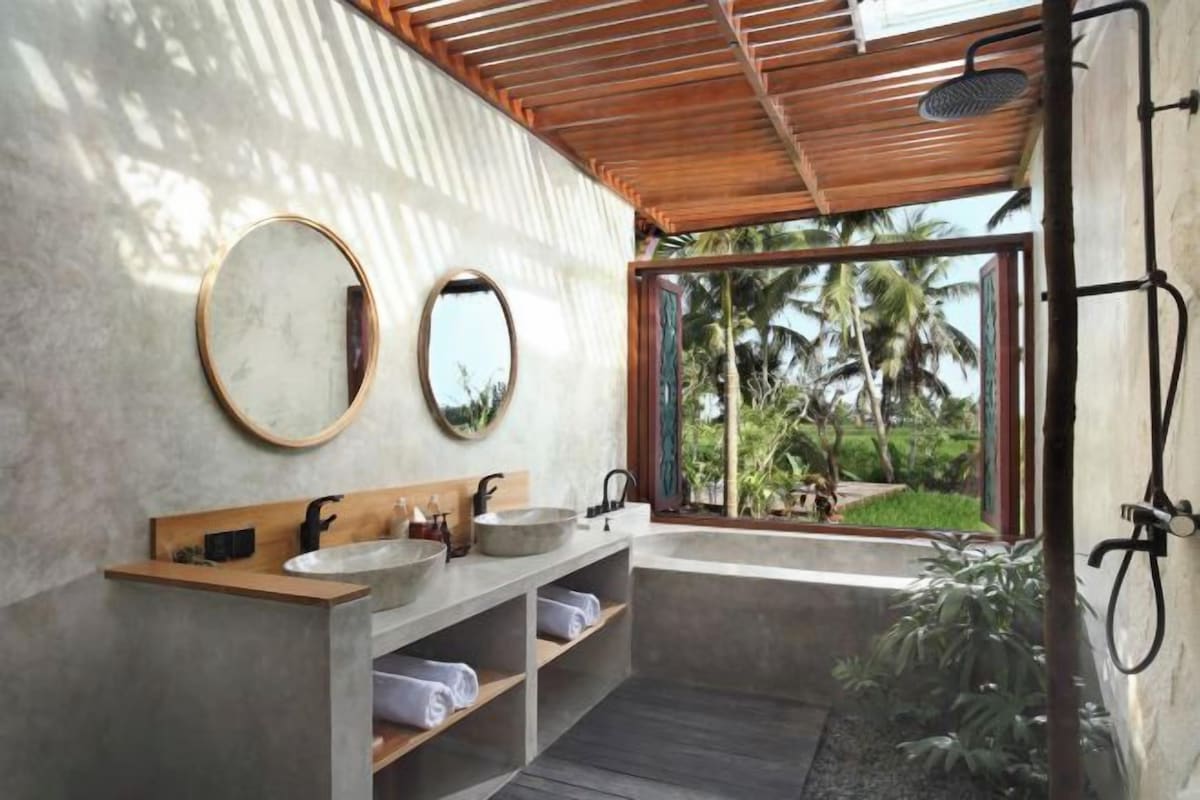 Un baño con dos lavamanos, ducha y una ventana grande con vistas a palmeras.