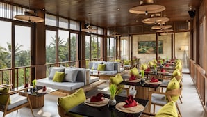 A spacious dining area with high ceilings and large windows, offering views of palm trees. Tables are set with green and red napkins, and there are cushioned seating areas. - Inara Alas Harum (Payangan)