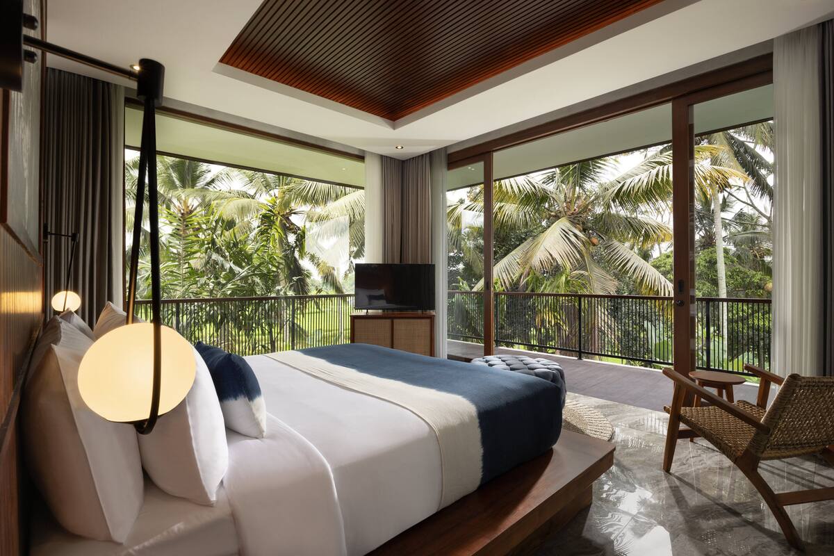 A spacious bedroom with a large bed, a TV, and a balcony with a view of palm trees.