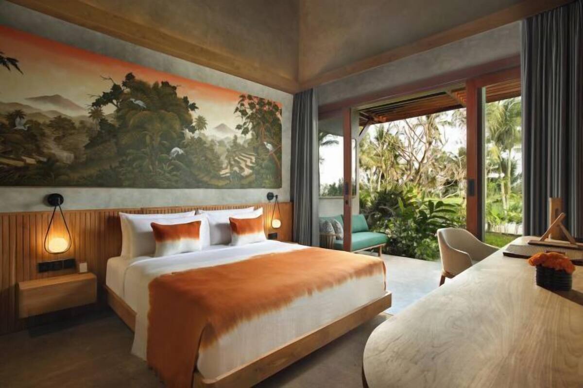 A modern hotel room with a large bed, a desk, and a view of greenery outside.