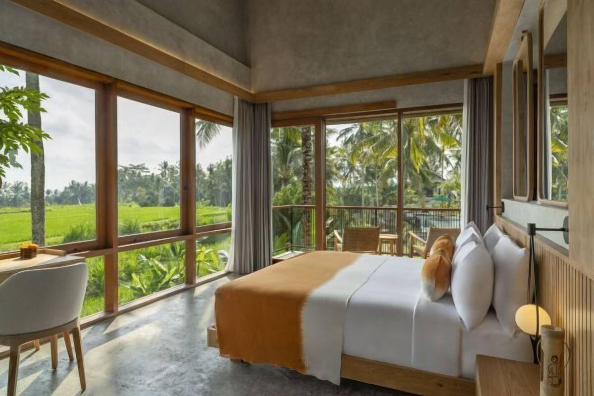A bedroom with a large bed, a desk, and a chair, with a view of a lush green landscape.