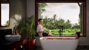 A bathroom with a large bathtub filled with flowers, a sink, and a window overlooking a lush garden. - Inara Alas Harum (Payangan)