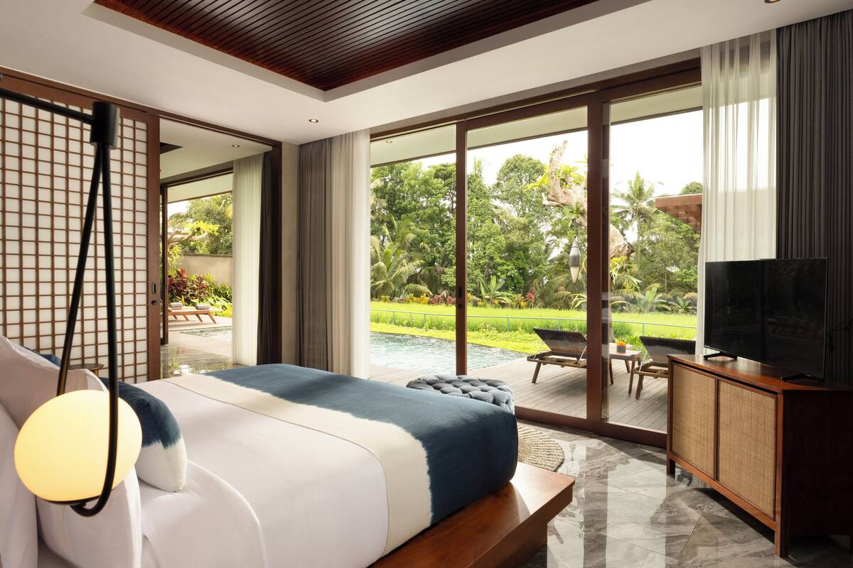 A modern hotel room with a large bed, a television, and a view of a garden and pool.