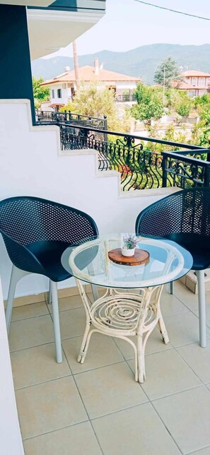 A balcony with two black chairs and a glass table, a decorative railing, and a view of houses and greenery in the background. - Vila Camellia  (Volvi)