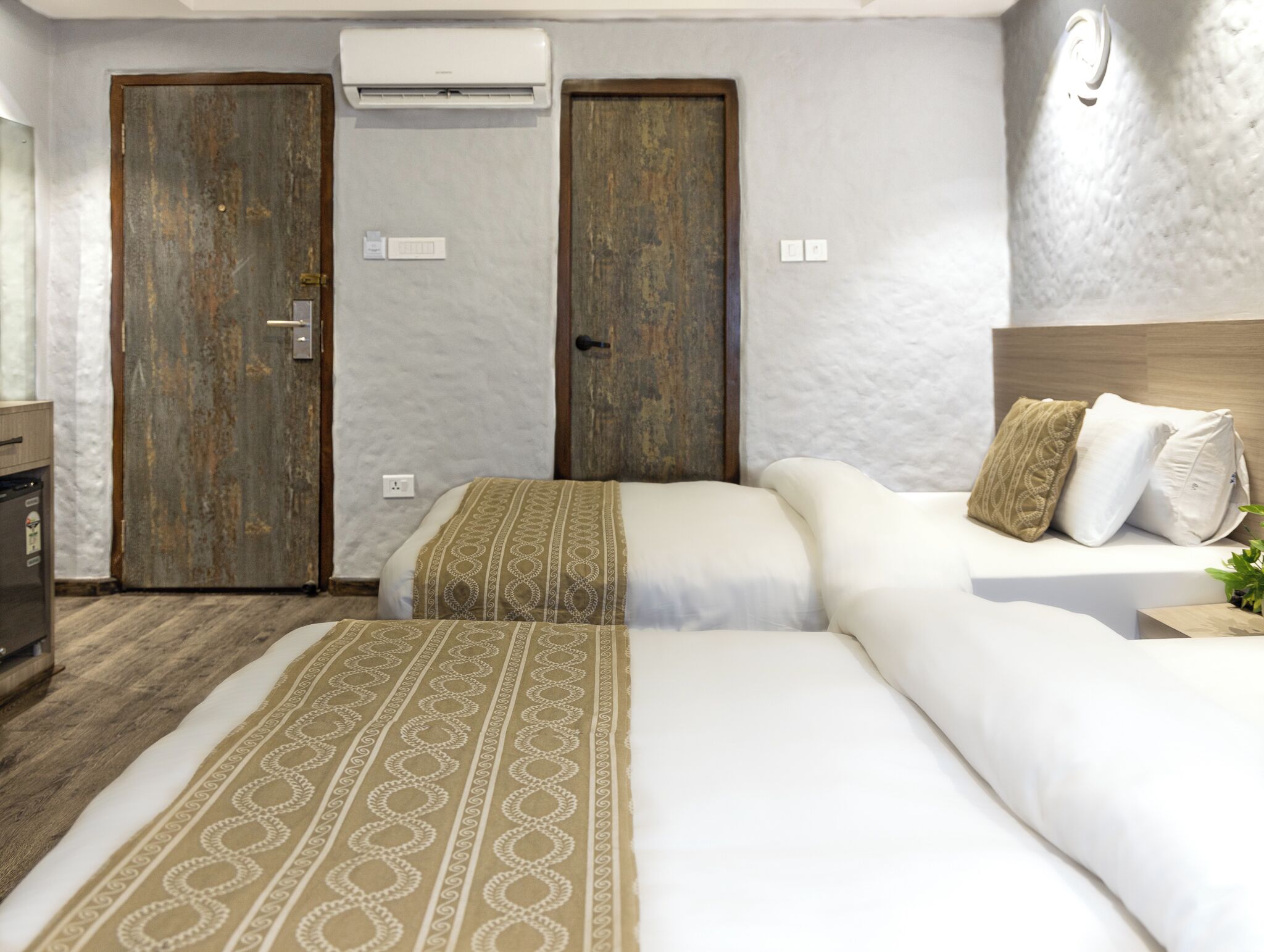 Habitación de hotel con dos puertas de madera, un equipo de aire acondicionado, una cama con ropa de cama estampada y una mesita de noche con una planta.