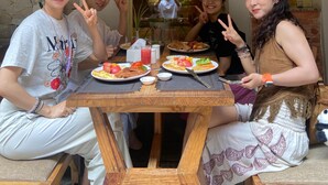 A group of people seated at a wooden table with plates of food, a stone wall, and a sign indicating a hotel. - Vastu Boutique Hotel (Kathmandu)