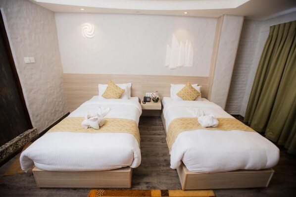 A hotel room with two beds, each with a white bedspread and a yellow accent pillow. There are towels on the beds, a nightstand with a phone and a vase of flowers, and a wall-mounted light fixture. - Vastu Boutique Hotel (Kathmandu)