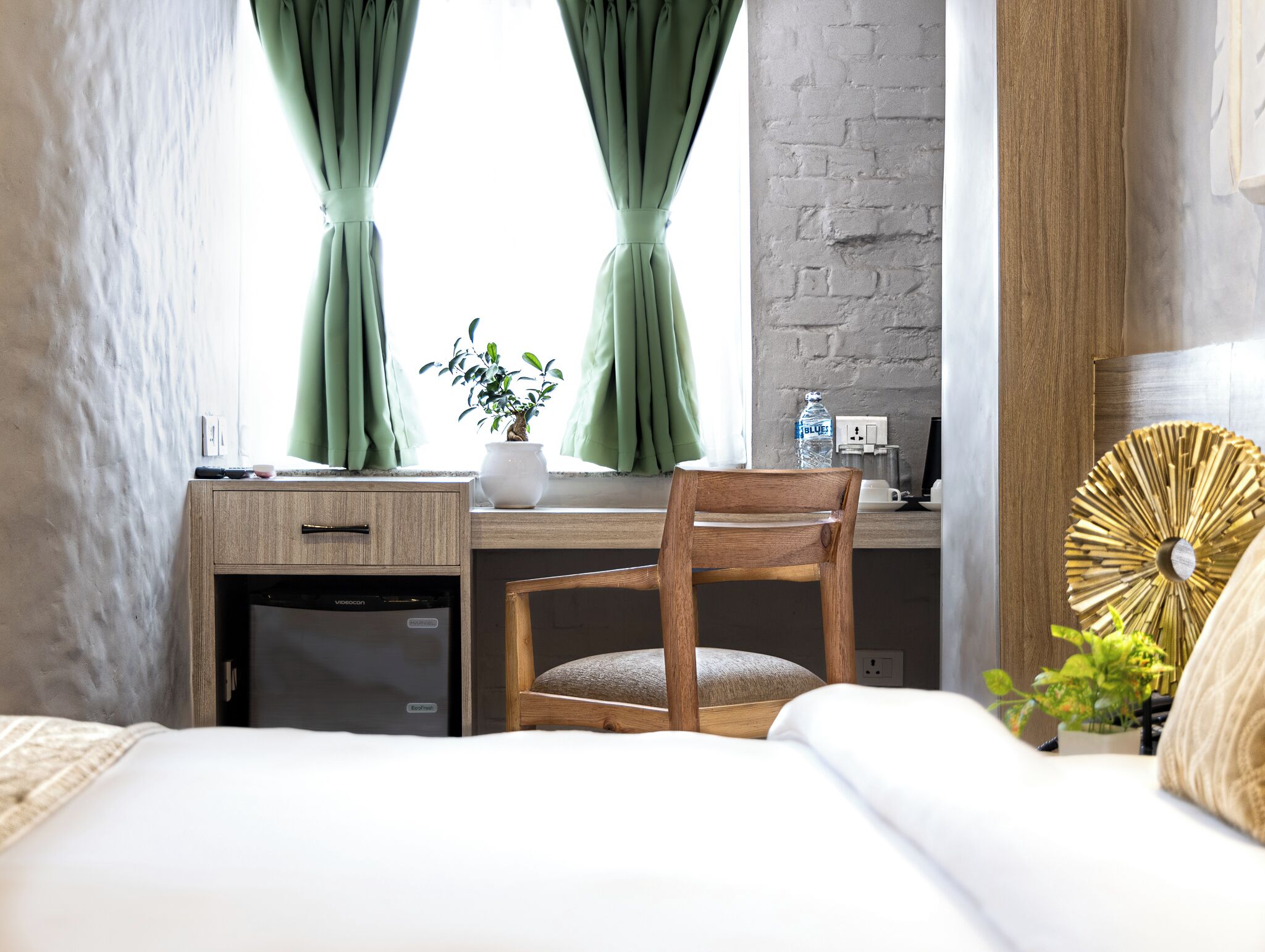 Un dormitorio con un escritorio de madera, una silla, una cama, una planta en maceta y un ventilador de pared.