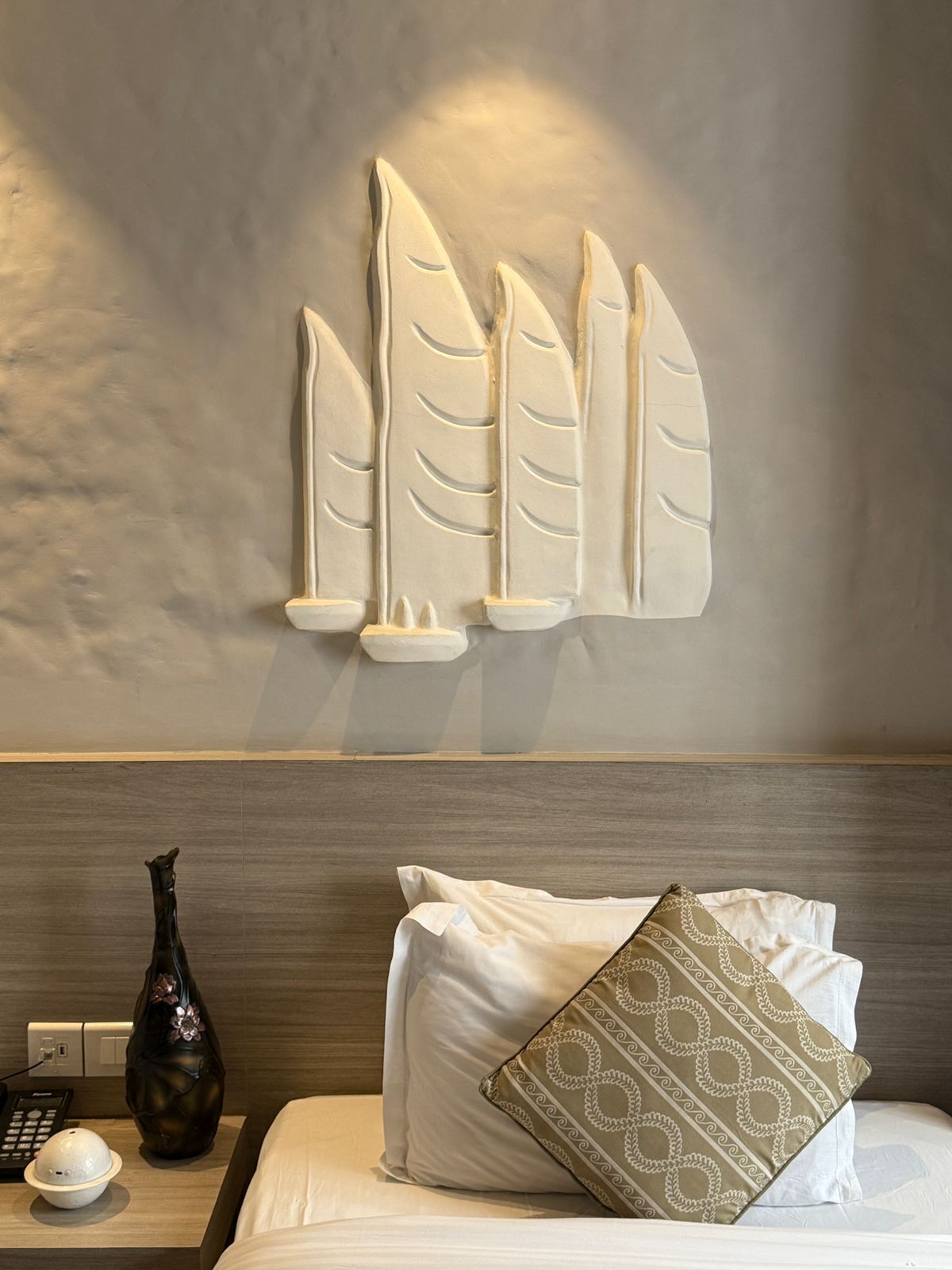 A hotel room with a bed, pillows, a vase, and a wall-mounted decorative piece.
