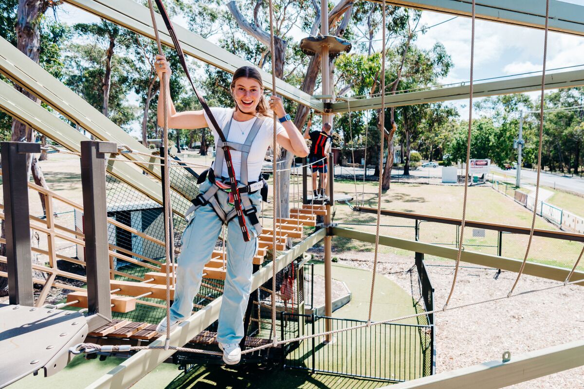 A person on a rope bridge with safety harnesses, surrounded by trees and a clear sky.