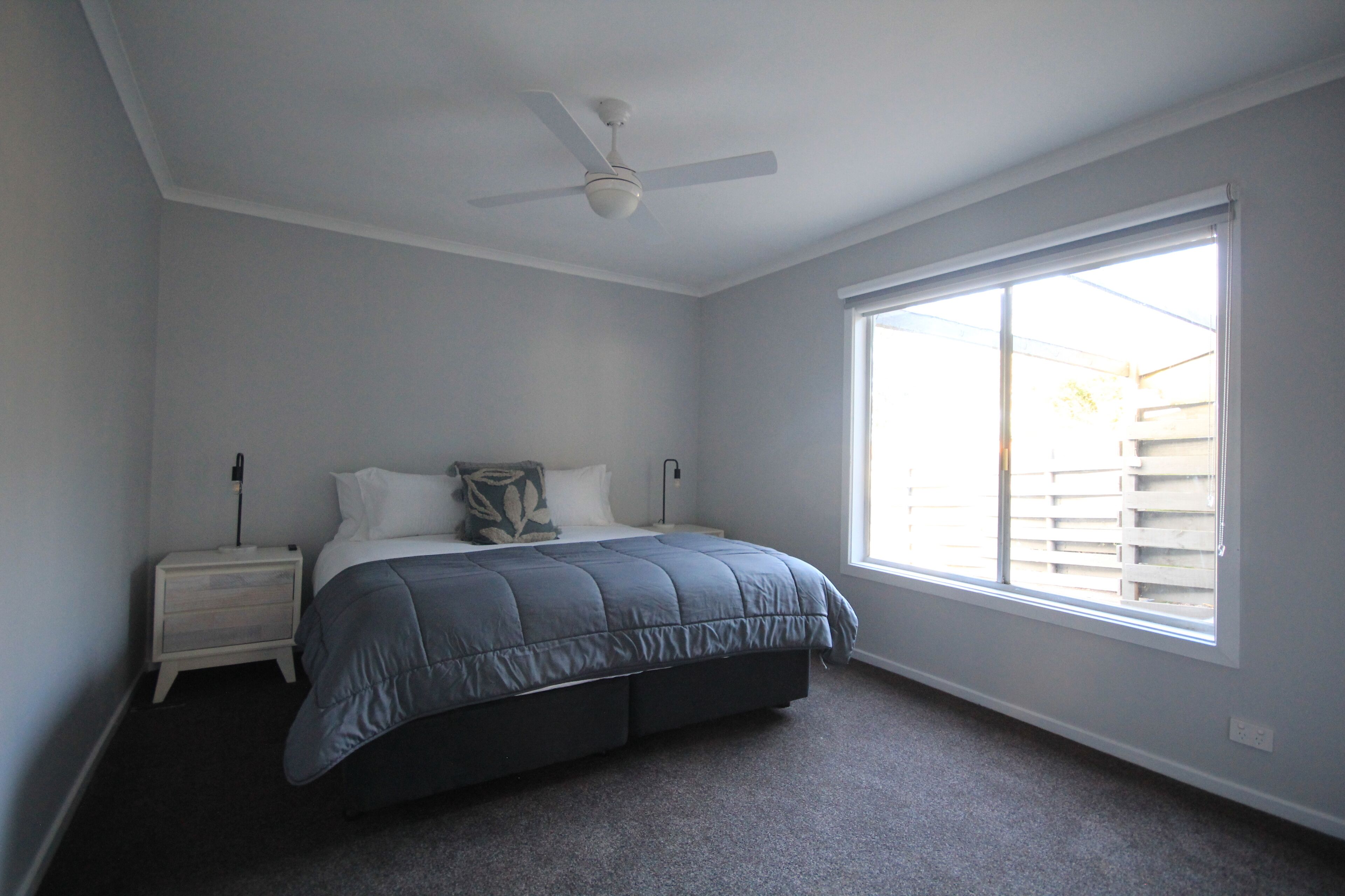 Une chambre à coucher avec un lit, une table de chevet, un ventilateur de plafond et une grande fenêtre.