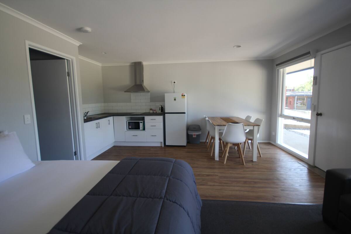 Una habitación compacta con cama, kitchenette, zona de comedor y un ventanal grande.