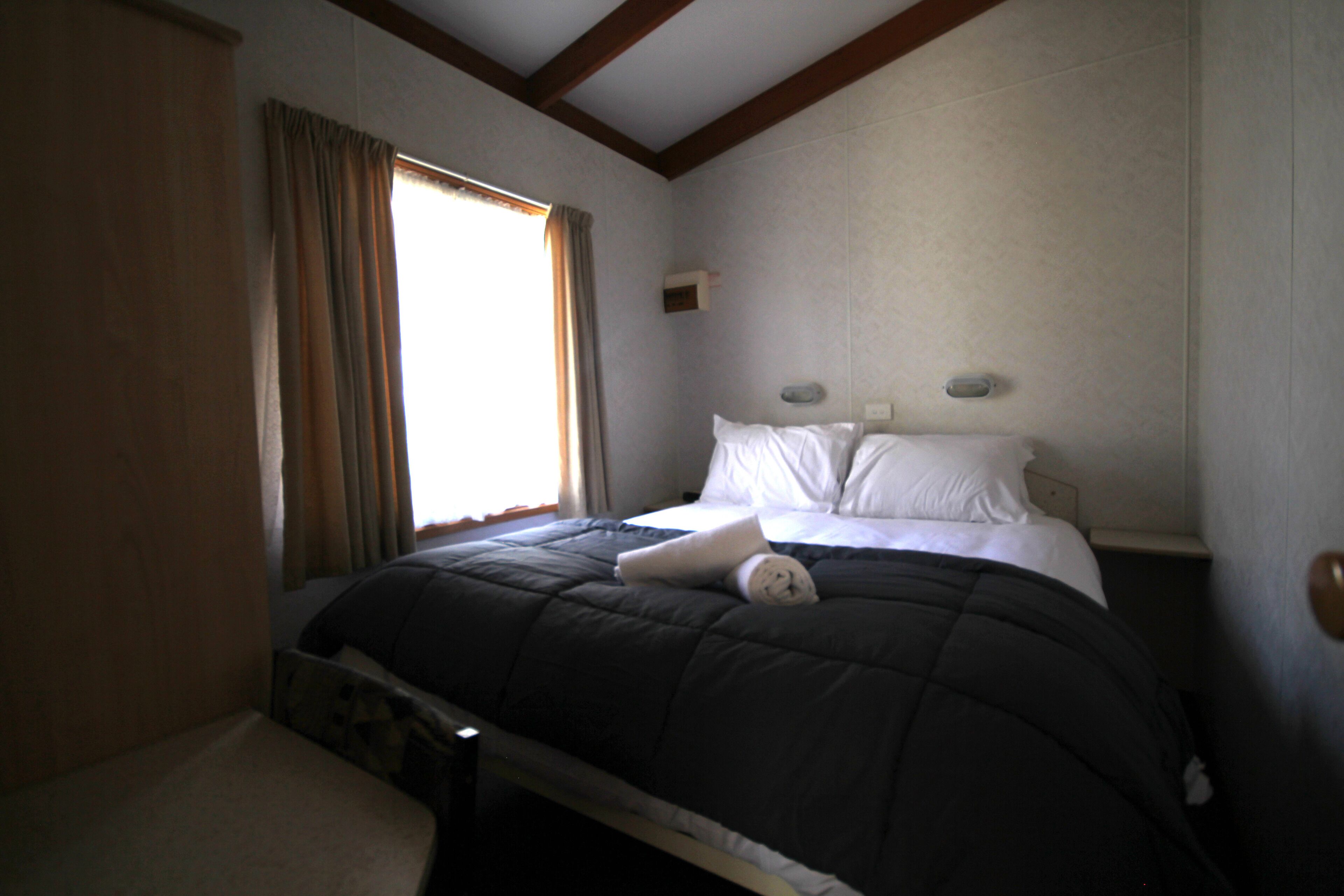 A bedroom with a bed, a window with curtains, a nightstand, and a wall-mounted light.