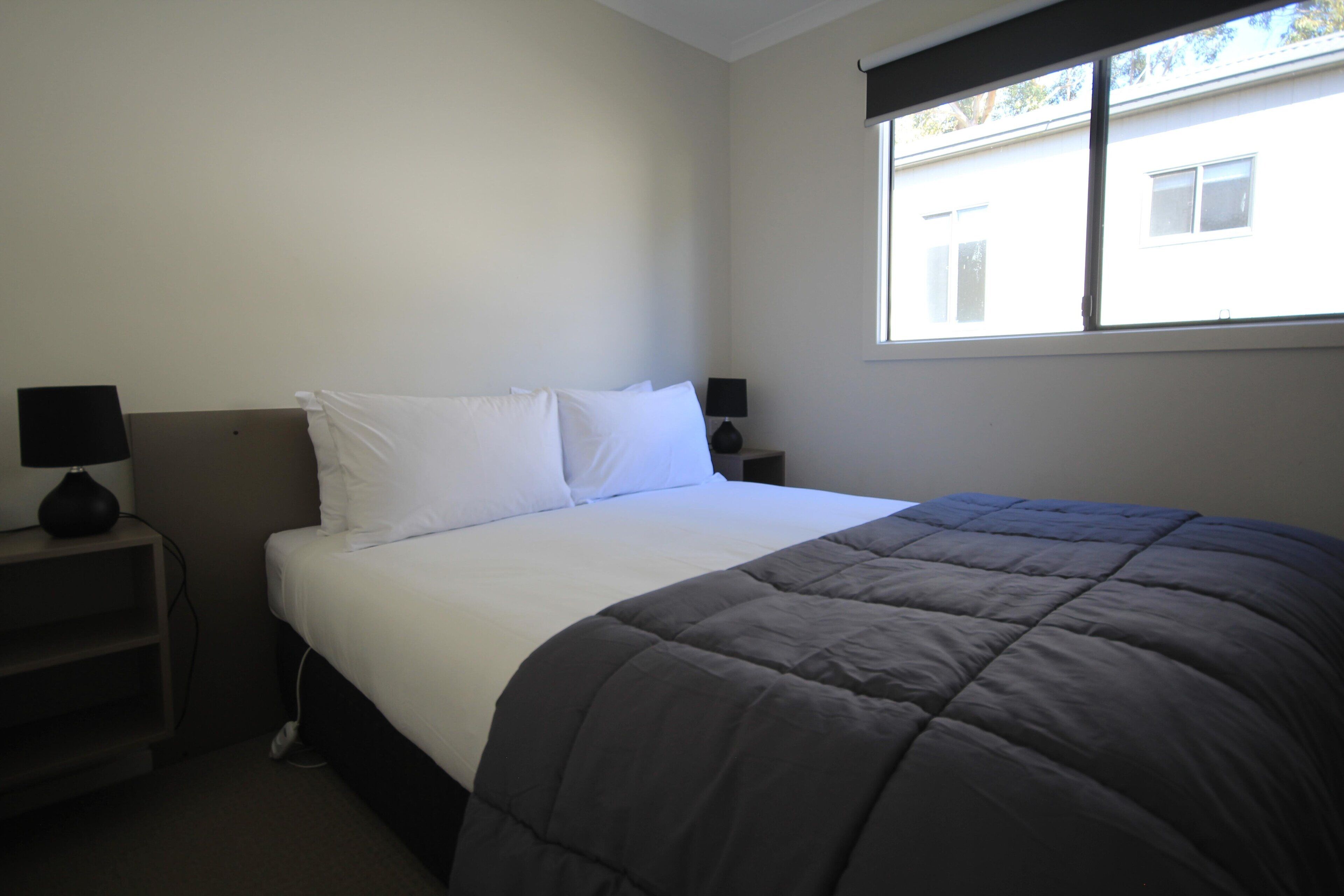 A neatly made bed with a dark blanket, two bedside tables with lamps, and a window with blinds.