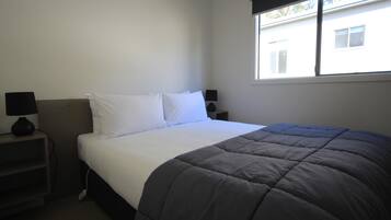 A neatly made bed with a dark blanket, two bedside tables with lamps, and a window with blinds.