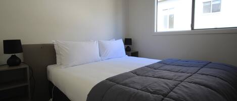 A neatly made bed with a dark blanket, two bedside tables with lamps, and a window with blinds.